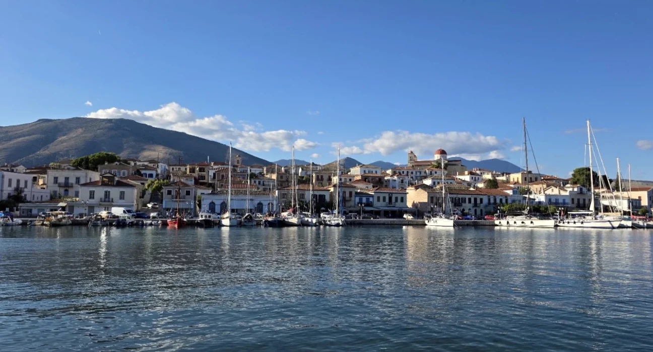 Panoramic view of Galaxidi’s historic harbour, with traditional waterfront houses, moored sailing boats, a red-domed church and mountains in the background.
