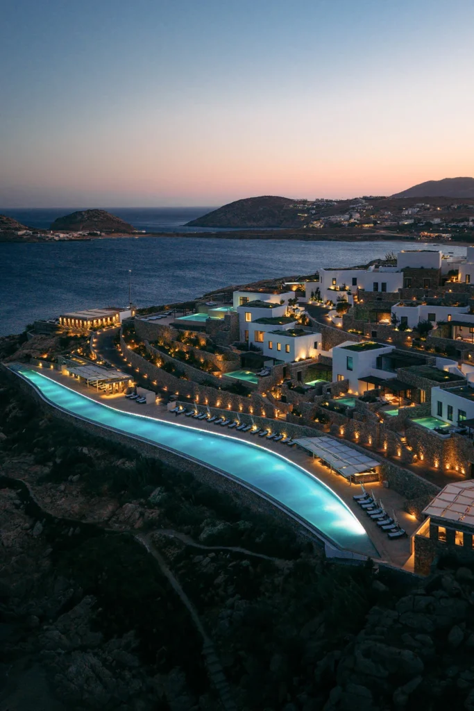 Cali Mykonos hotel and its illuminated infinity pool overlooking the Aegean Sea at dusk.