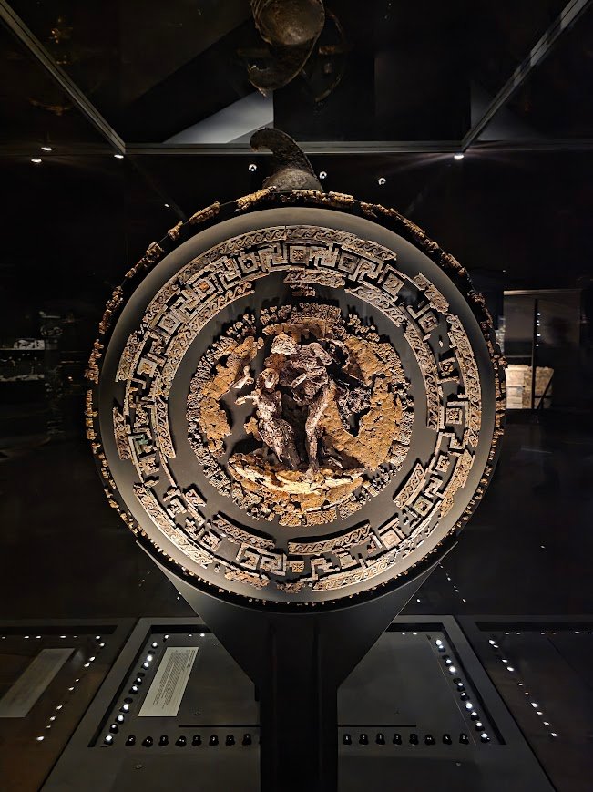 The gold and ivory shield of Philip II, featuring a central depiction of Achilles defeating the Amazon queen Penthesileia. Constructed from layered wood, leather, and cords, reinforced with gilded silver plates and detailed reliefs of lions, winged Victories, and Heracles’ club. The interior is coated in gilded stucco with ivory meanders and cast-glass inlays. Museum of the Royal Tombs of Aigai, Expats Athens, Expats Greece, Xpat.gr