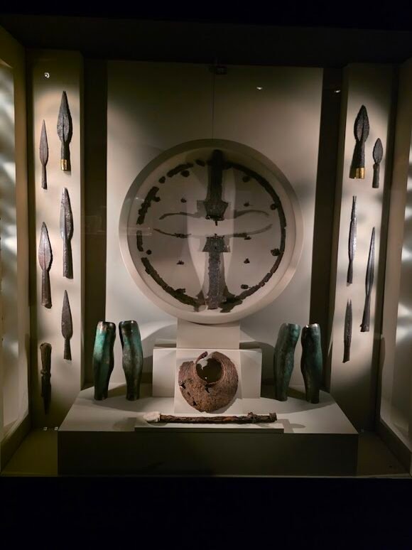 Weaponry of Philip II (336 BCE): Philip’s weapons and armor highlight the dual role of arms in Macedonian society—as essential tools in combat and as prestigious symbols of status. While typical burials in Aigai included two spears or javelins, occasionally a sword, and for the wealthiest, a helmet, Alexander the Great exceeded tradition for his father’s funeral, providing four complete suits of armor: one for the pyre and three as grave goods, all expertly crafted and trimmed with gold. Additional finds from the tomb include a wood, leather, and cloth shield reinforced with iron and decorated with embossed lions; a pectoral with iron plate; an iron sword with ivory-inlaid wooden sheath; two pairs of bronze greaves; an iron sarissa head; an iron spike for anchoring the spear; and nine iron spear and javelin heads, two with broad golden plates. Museum of the Royal Tombs of Aigai, Expats Athens, Expats Greece, Xpat.gr