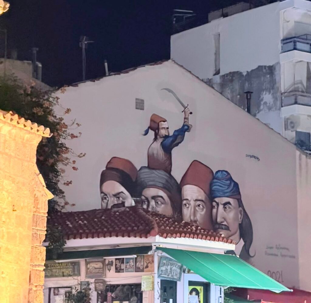 A mural by Skitsofrenis in 23rd March Square, in Kalamata’s historic centre, commemorating the Greek War of Independence. Created in March 2021, it portrays Papaflessas, Nikitaras, Petrobey Mavromichalis and Theodoros Kolokotronis. Image credit: Xpat.gr.