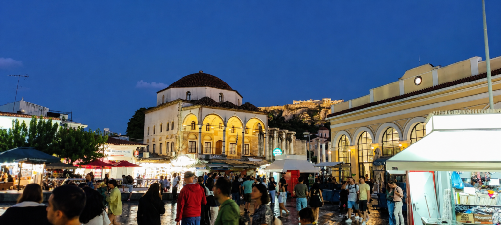Evening crowds in Monastiraki Square with the Tzistarakis Mosque and illuminated Acropolis in the background.