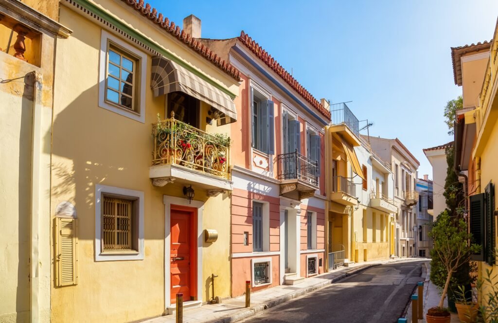 Quiet residential street in Plaka, Athens, with pastel houses, shaded balconies and closed shutters on a summer afternoon.
