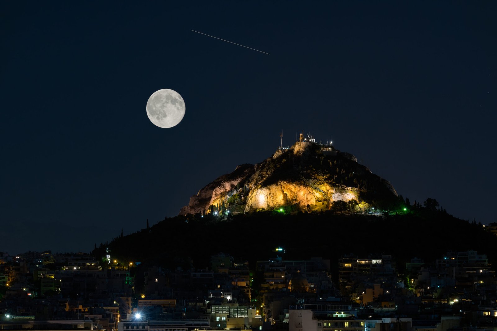 Lycabettus Hill in Athens silhouetted against the August full moon, with a meteor streaking across the night sky.