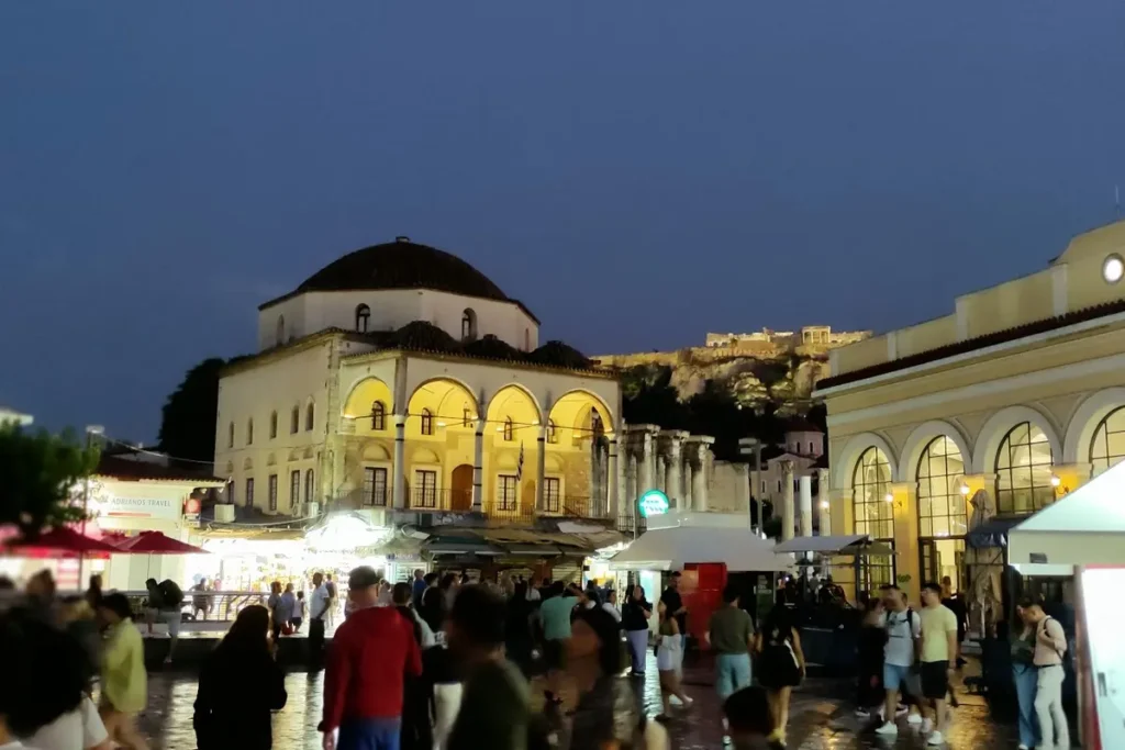 Evening crowds in Monastiraki Square with the Tzistarakis Mosque and illuminated Acropolis in the background.