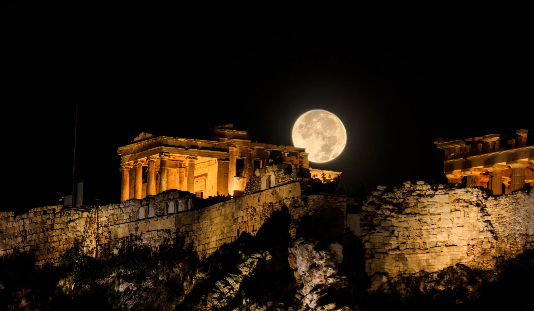 Full moon rising above the illuminated Parthenon and ancient walls of the Acropolis in Athens at night.