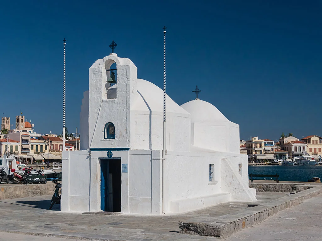 Whitewashed Agios Nikolaos chapel with a bell tower and blue doorway beside Aegina’s harbour.
