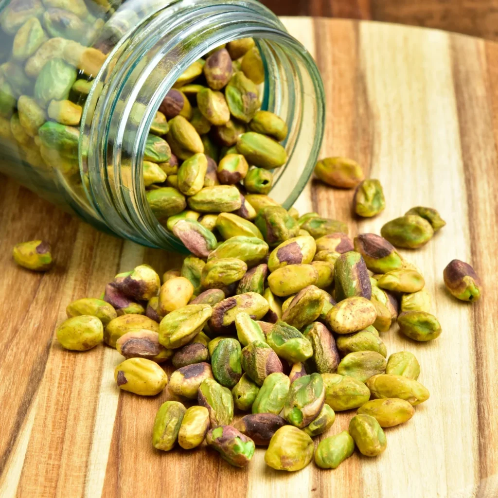 Shelled pistachio kernels spilling from a glass jar onto a wooden surface.
