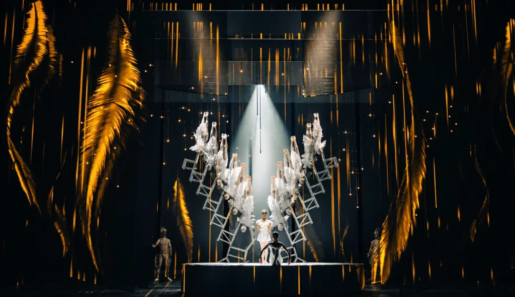 SWAN performers create a symmetrical acrobatic formation beneath a spotlight and golden stage projections.