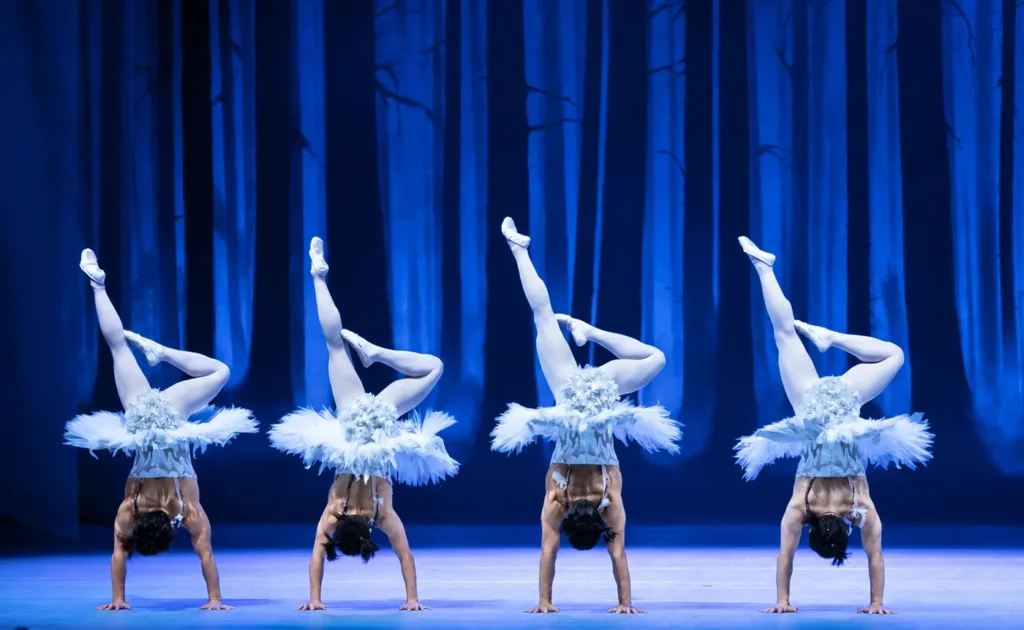 Four SWAN acrobats perform synchronised inverted balances against a blue forest backdrop.