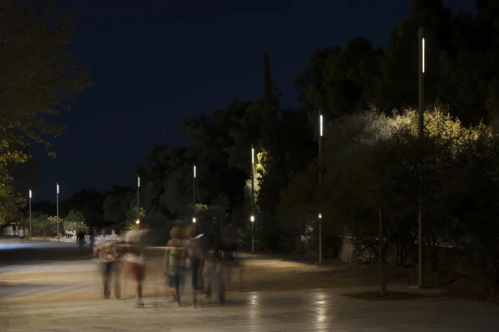 Pedestrians walking beneath a row of new lighting poles along Athens’ historic promenade at night